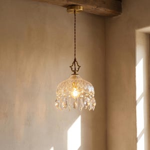 Può includere: Una lampada a sospensione in stile vintage con un paralume in vetro trasparente e decorato e pendenti di cristallo. Il lampadario ha un baldacchino e elementi decorativi color ottone. La lampadina è visibile, emettendo una calda luce.
