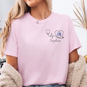 May include: A pale pink t-shirt with a stethoscope design, a heart containing the letters "RN", and the name "Sophia" printed below. The shirt is made of a soft material.