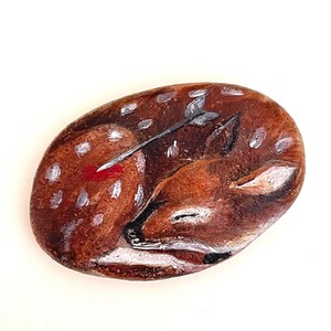May include: Hand-painted rock art depicting a sleeping fawn with an arrow through its side. The fawn is brown with white spots, and the arrow has a white shaft and grey arrowhead. The oval-shaped rock has a brown background.