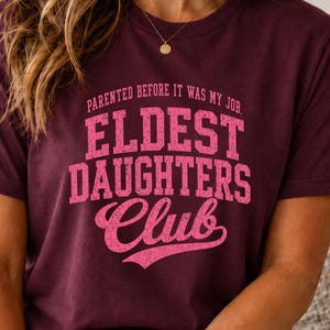 May include: A maroon short-sleeved t-shirt featuring the pink text "PARENTED BEFORE IT WAS MY JOB. ELDEST DAUGHTERS CLUB" printed on the front in a distressed, collegiate-style font with a script accent for the word Club.