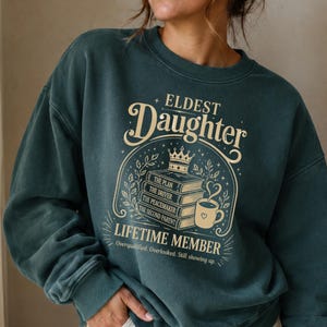 May include: A dark teal crewneck sweatshirt featuring a cream-colored graphic with the text "Eldest Daughter Lifetime Member" above an illustration of a stack of books topped with a crown and a steaming mug, with smaller text below the graphic.