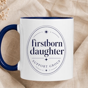 May include: A white ceramic mug with a dark blue handle and interior. The front features a dark blue oval emblem with the text "firstborn daughter" and "SUPPORT GROUP" below it, accented by small star symbols at the top and bottom of the design.