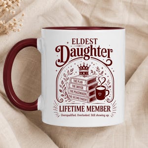 May include: White ceramic mug with a burgundy interior, handle, and text. The mug features the words "ELDEST Daughter" in a decorative font, along with the text "LIFETIME MEMBER" and the phrases "Overqualified. Overlooked. Still showing up."