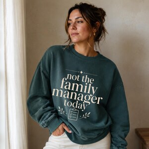 May include: A dark teal crewneck sweatshirt featuring the cream-colored text "not the family manager today" printed on the front. The design includes a small star icon above the text and a clipboard icon with a checkmark flanked by leaves below.