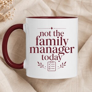 May include: A white ceramic mug with a dark red handle and interior rim. The front features dark red text that reads "not the family manager today" above a small graphic of a checklist on a clipboard, flanked by two simple leaf sprigs.