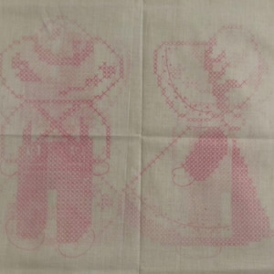 May include: A piece of off-white fabric featuring a pink cross-stitch pattern of two figures. The left figure wears a hat and overalls, while the right figure wears a bonnet and a dress. The design is printed in a light pink ink on the textile.