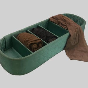 May include: An elongated, quilted green storage organizer with four internal compartments. The organizer contains folded hosiery in shades of brown and black, with a sheer brown fabric piece draped over the outer edge of the container.