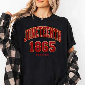Può includere: Una t-shirt nera con la scritta JUNETEENTH in un carattere universitario arcuato rosso e giallo, il numero 1865 in grandi lettere rosse e gialle e la parola FREEDOM in lettere rosse più piccole posizionate nella parte inferiore.