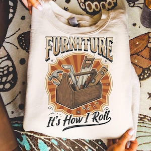 May include: A cream-coloured sweatshirt with a graphic of a wooden toolbox filled with tools, including a hammer, saw, and level. The word "FURNITURE" is arched above the toolbox, with the phrase "It's How I Roll" below.