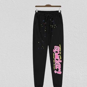 May include: Black sweatpants with a drawstring waist and pockets. The pants feature colorful splatter details and the text "powder!" in pink and yellow, with additional text in a script font.