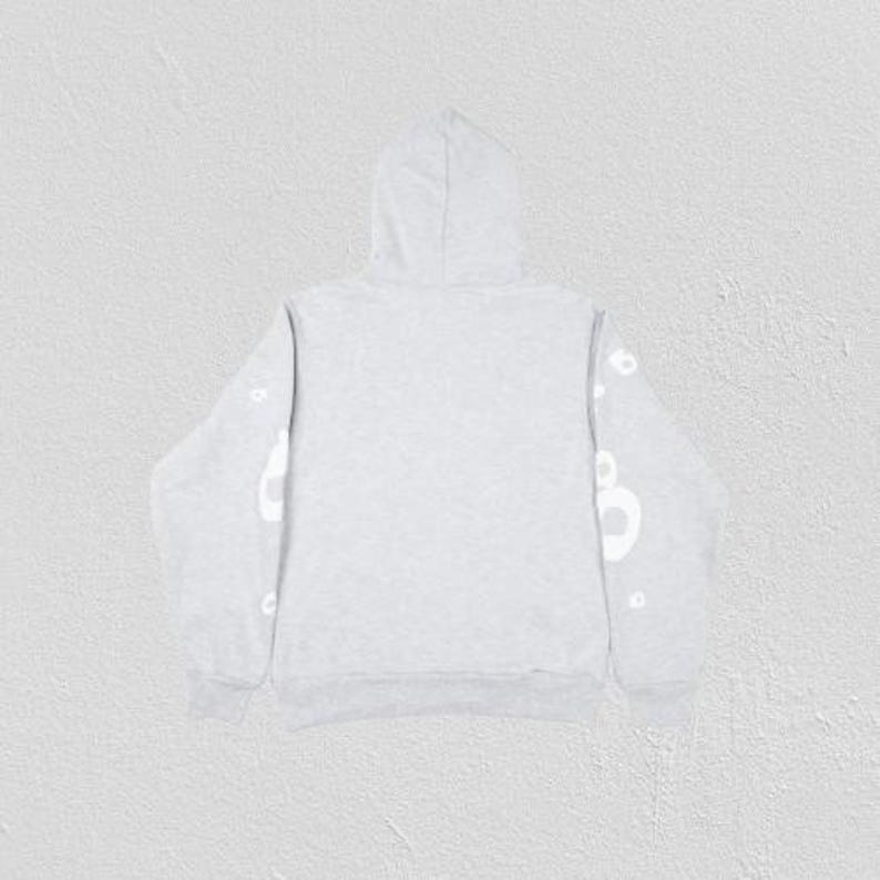 May include: A light gray hoodie with a hood and long sleeves. White lettering is visible on the sleeves. The hoodie is made of a soft material and is suitable for casual wear.