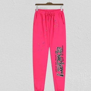 May include: Bright pink sweatpants with a drawstring waist and pockets. The pants feature a graphic on the lower leg and colorful paint splatters. The graphic reads "Somebody" in a stylized font.