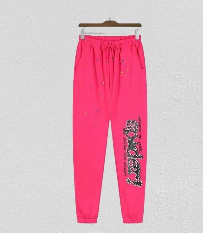 May include: Bright pink sweatpants with a drawstring waist and pockets. The pants feature a graphic on the lower leg and colorful paint splatters. The graphic reads "Somebody" in a stylized font.