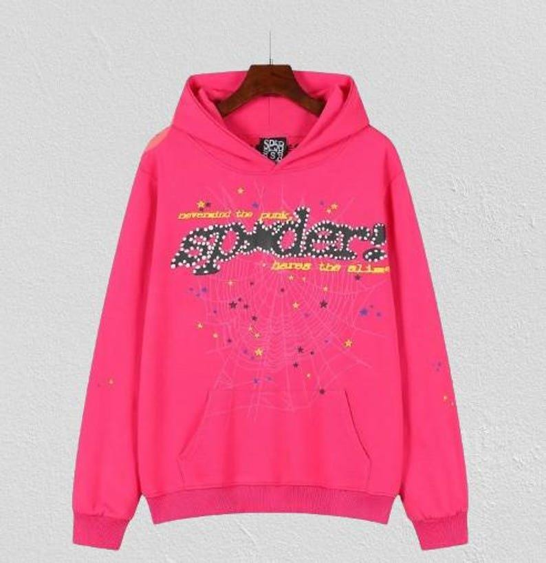 May include: A bright pink hoodie with a front pocket and a drawstring hood. The front features the word "Spider" in a stylized font, with a spiderweb design and the text "defended the punk" and "bares the alibi".