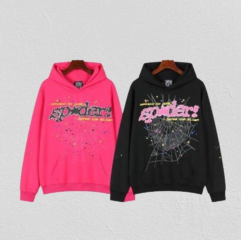 May include: Two hooded sweatshirts, one hot pink and one black, each featuring a spider web design. The word "spider" is prominently displayed in a stylized font across the chest of each hoodie. The sweatshirts have a front pocket and a drawstring hood.
