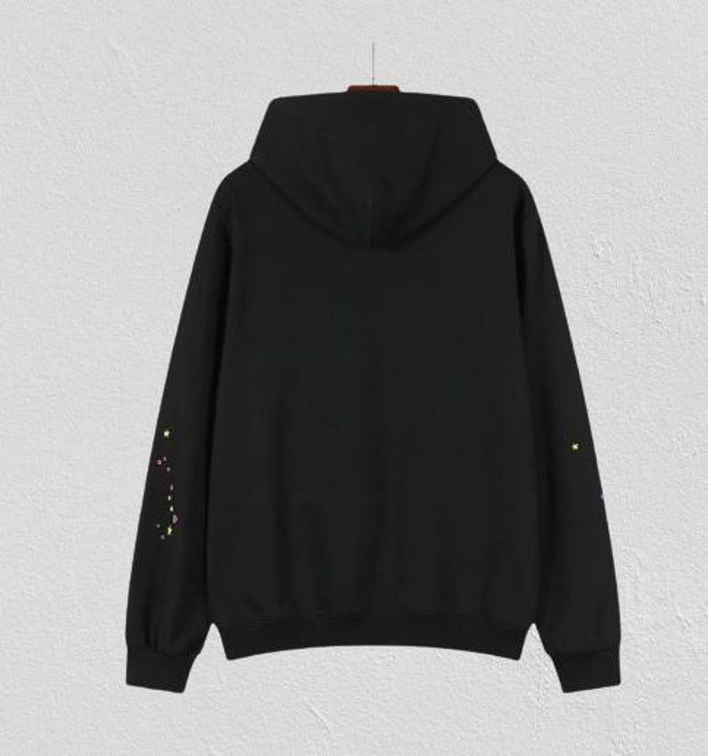 May include: A black hoodie with a hood and long sleeves. The sleeves feature a constellation design in yellow and white. The hoodie is hanging on a hanger against a white textured wall.
