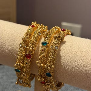 Peut inclure: Deux bracelets rigides dorés ornés de filigrane métallique complexe et de petites breloques pendantes. Les bracelets sont agrémentés de pierres cabochons ovales rouges et vertes alternées, fixées le long de la surface métallique texturée.