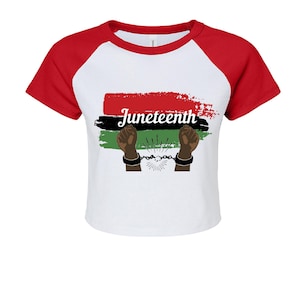 May include: A white raglan t-shirt with red sleeves featuring a graphic of red, black, and green brushstrokes, the word Juneteenth in white script, and two dark-toned raised fists with broken chains on their wrists centred on the front.