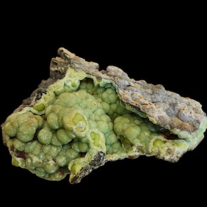 May include: A raw mineral specimen featuring a cluster of pale green, botryoidal formations nestled within a rough, dark gray, and textured rocky exterior. The interior displays smooth, rounded, bubble-like structures in varying shades of light green.