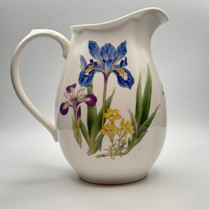 May include: A white ceramic pitcher featuring a botanical illustration of blue and purple irises alongside small yellow flowers and green leaves. The pitcher has a curved handle and a wide pouring spout, with a smooth, glossy finish throughout.