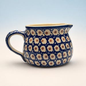 May include: A ceramic creamer pitcher featuring a rounded body and a curved handle. The exterior is decorated with a repeating pattern of small, dark blue floral shapes with white petals and brown centers against a cream-colored background.
