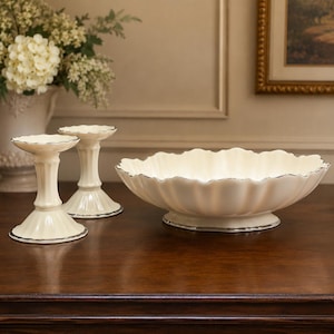 May include: A set of two cream-colored ceramic candle holders and a matching shallow bowl, each featuring a scalloped edge and a thin silver metallic trim along the rim and base, displayed together on a dark wooden surface.