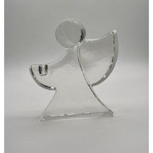 Op de afbeelding: Een kandelaar van helder glas in de vorm van een abstracte engelfiguur. Het ontwerp heeft een cirkelvormig hoofd, een breed uitlopend lichaam en één vleugel die naar rechts uitsteekt. Aan de linkerkant is een kleine cilindrische kaarsenhouder verwerkt.