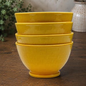 May include: A stack of four bright yellow ceramic bowls featuring a raised basketweave texture on the exterior. Each bowl has a rounded shape with a flared rim and a small circular pedestal base, creating a uniform set of kitchenware.