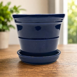 Puede incluir: Una maceta de cerámica de color azul marino profundo con acabado brillante, que presenta un borde ensanchado y una base de platillo circular a juego. La maceta tiene una superficie lisa y sólida con sutiles crestas horizontales en el exterior.