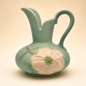 May include: A light green ceramic pitcher with a crackle glaze finish. The pitcher features a prominent embossed white flower with a yellow and green center on the front, along with a slender handle and a curved pouring spout.