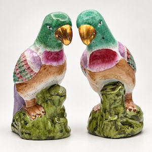 May include: A pair of ceramic parrot figurines perched on green textured bases. Each bird features a vibrant green head, a metallic gold beak, a magenta chest, and wings with detailed green and pink feather patterns over a tan body.