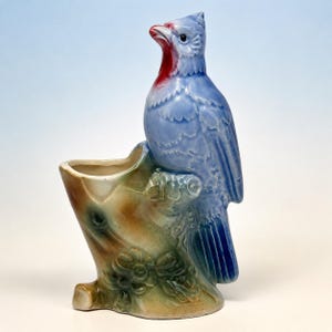 May include: A ceramic planter shaped like a blue bird perched on a brown and green tree stump. The bird has a red throat patch and a glossy finish. The stump features moulded floral details at the base and a hollow opening for small plants.