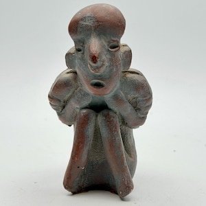 May include: A small, hand-sculpted terracotta figurine depicting a seated figure with a prominent, rounded head, stylised facial features, and arms resting on raised knees. The surface has a weathered, matte finish with reddish-brown and muted grey tones.