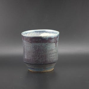 May include: A ceramic cup featuring a mottled glaze in shades of deep blue, purple, and grey. The exterior surface displays horizontal ridges near the base, while the rim shows a lighter, cream-colored glaze drip against the darker body tones.