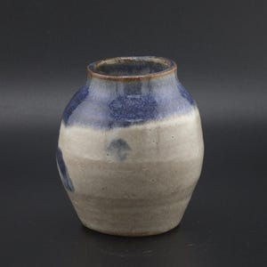 May include: A small, rounded ceramic vase featuring a two-tone glaze. The upper portion is a deep, saturated blue that transitions into a light, speckled beige on the lower body. The rim has a thin, exposed brown clay edge with a smooth texture.