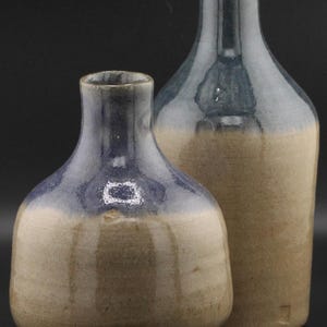 May include: Two ceramic vases with a two-tone design. The top portion of each vase is a deep blue, transitioning to a tan base. The vases have a glossy finish and are set against a black background. One vase is shorter and rounder, while the other is taller and more cylindrical.