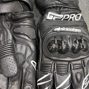Könnte beinhalten: Schwarze Motorradhandschuhe aus Leder mit weißen Akzenten. Die Handschuhe verfügen über Knöchelschutz und den Text "GP PRO" und "Alpinestars". Die Handschuhe haben eine strukturierte Oberfläche und sind für das Motorradfahren konzipiert.