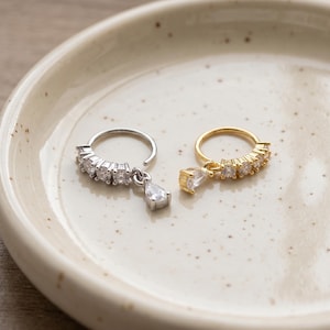 Puede incluir: Dos anillos circulares, uno plateado y otro dorado, cada uno con una hilera de cuatro pequeñas piedras redondas transparentes y un dije colgante en forma de pera. Las piezas de joyería se muestran juntas sobre una superficie lisa.