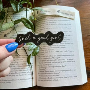 May include: A black sticker with the phrase "such a good girl" in a cursive font. The sticker is held by a hand with blue nail polish, with an open book and greenery in the background.