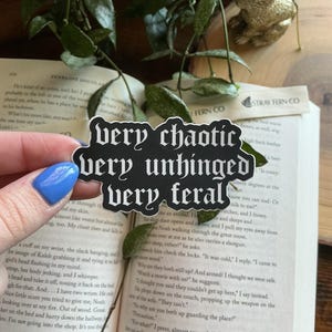 May include: A black and white sticker with the words "very chaotic, very unhinged, very feral" in a gothic font. The sticker is held over an open book with text and a sprig of greenery. A golden rabbit figurine is in the background.