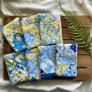 May include: A collection of six rectangular fabric pouches featuring a quilted patchwork design in shades of yellow, white, and blue. The pouches display various floral and paisley patterns, with visible stitching lines and white zipper closures.