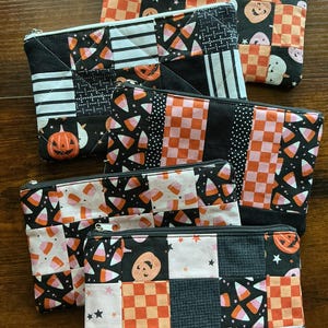 May include: A collection of five rectangular zippered pouches featuring patchwork designs with Halloween-themed patterns including orange and black checkered squares, candy corn prints, smiling jack-o'-lanterns, and small black stars on white fabric.