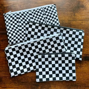 May include: A collection of four rectangular zippered pouches in varying sizes, each featuring a consistent black and white checkered pattern. The pouches are constructed from fabric and include a metal zipper closure along the top edge of each item.