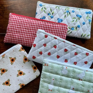 May include: Five rectangular zippered pouches featuring various fabric patterns: a white quilted pouch with blue blueberries, a red and white gingham pouch, a white quilted pouch with red strawberries, a white pouch with sunflowers, and a green gingham pouch with white geese.