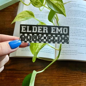 May include: A rectangular sticker with a black and white checkered border and the text "ELDER EMO BOOK CLUB" in a bold, white, collegiate-style font. The sticker is positioned in front of an open book and a green and white variegated plant.