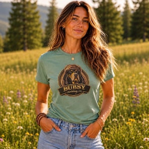 May include: A sage green short-sleeved t-shirt featuring a circular graphic of a dog wearing a hat with the text BUBSY clothing, Co. printed in a dark brown and gold emblem style on the chest, paired with light blue denim jeans.