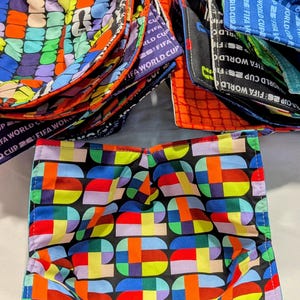Puede incluir: Una colección de soportes de tela para cuencos con un vibrante patrón geométrico multicolor y el texto FIFA WORLD CUP 26. Los soportes cuadrados y acolchados están apilados, mostrando varias combinaciones de colores, incluyendo rojo, azul y verde.