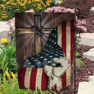 May include: A wooden cross with a red, white, and blue American flag draped over it. The flag is partially covering the cross, and the hand of a person is visible in the lower right corner of the image. The background is a wooden plank.