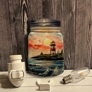 Puede incluir: Un tarro de vidrio pintado a mano con una escena de faro. El tarro tiene un cielo de puesta de sol con un cielo rojo y naranja, un faro con un techo rojo y un océano azul con olas. El tarro es una luz nocturna con una base blanca enchufable.