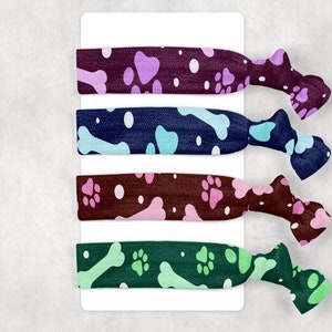 May include: Four colorful hair ties with dog-themed patterns. Each tie features a different design: paw prints and bones in shades of purple, blue, brown, and green. The ties are attached to a white card, perfect for pet lovers.